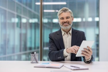 Modern bir ofis masasında tablet kullanan kendinden emin orta yaşlı bir profesyonel. Düşüncelilik, teknoloji kullanımı ve işyeri ortamını resmediyor, profesyonelliği aktarıyor