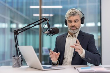 Kulaklık takan, mikrofona konuşan, modern ofis ortamında dizüstü bilgisayarla oturan profesyonel bir yayıncı. Bireyler açık bir iletişim gösteriyor.