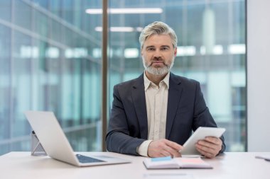 Gri saçlı profesyonel bir işadamı, modern ofis ortamında cam duvarlarla çevrili bir masada otururken teknolojiyle etkileşime girer..