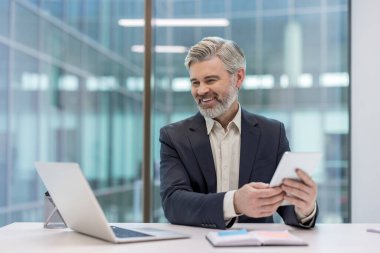 Modern bir ofiste kendine güvenen bir işadamı elinde tabletle gülümsüyor..