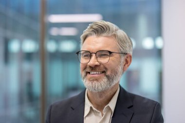 Sakallı, mutlu, orta yaşlı bir adam parlak bir ofiste gülümsüyor. Modern arka plan ve olumlu ifade, başarıyı resmetmek için ideal sıcaklık, güven ve profesyonelliği iletir.