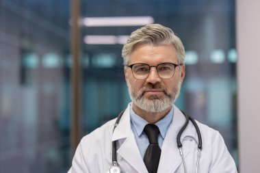 Modern bir sağlık merkezinde gözlük ve steteskop takan deneyimli bir erkek doktor profesyonel olarak ayakta duruyor. Sağlık hizmetleri ve tıbbi uzmanlık konuları için ideal