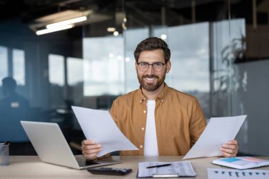 Evrak işlerini incelerken gülümseyen mutlu bir işadamı ve modern bir ofiste bilgisayarla çalışırken üretkenlik ve iş memnuniyetini gösteriyor.