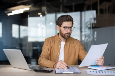 Gözlük takan, finansal belgeleri analiz eden ve modern ofiste masa başında oturup grafikler ve çizelgeler üzerinde çalışırken dizüstü bilgisayar kullanan odaklı iş adamı.