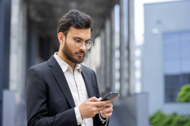 Takım elbiseli ve gözlüklü genç işadamı, bir şehir ortamında akıllı telefon kullanıyor, internette geziniyor ve en son gelişmeleri kontrol ediyor.