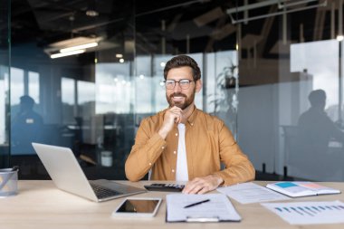 Mutlu muhasebeci finansal belgelerle çalışıyor, dizüstü bilgisayar ve hesap makinesi kullanıyor, satış çizelgelerini analiz ediyor, modern ofiste masa başında oturuyor, çenesine dokunuyor ve gülümsüyor.