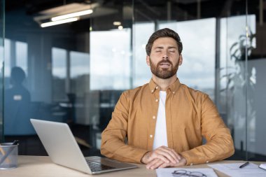 Ofis çalışanı iş yerinde farkındalık pratiği yapıyor, kapalı gözlerle derin bir nefes alıyor, dizüstü bilgisayarla ve belgelerle masada oturuyor, kurumsal çevrede zihinsel sağlık ve stres azaltmayı destekliyor.