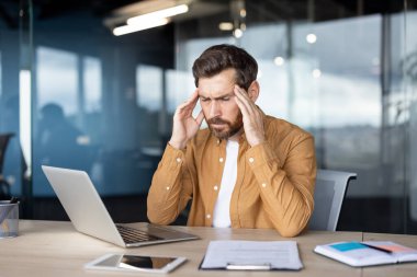 İşadamı, iş yerinde dizüstü bilgisayar ve belgelerle otururken baş ağrısı çekiyor ve tapınaklara dokunuyor. Stres ve aşırı çalışmadan muzdarip.