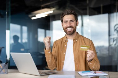 Neşeli işadamı, kredi kartını kullanarak, modern e-ticaretin kolaylığını ve kolaylığını vurgulayarak, başarılı bir online alışverişin ardından heyecanını dile getiriyor.