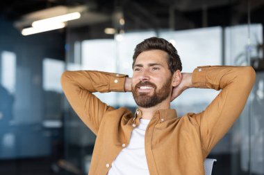 Gülümseyen iş adamı, elleri başının arkasında rahat bir şekilde koltuğunda oturuyor, modern ofiste huzur ve sessizliğin tadını çıkarıyor.