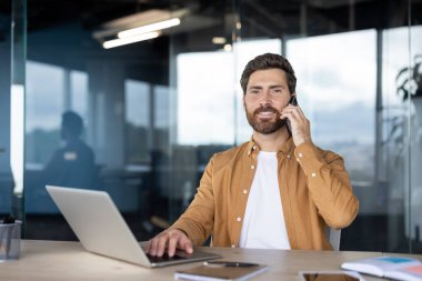 Gülümseyen iş adamı modern ofis masasında birden fazla iş yapıyor, bir dizüstü bilgisayar üzerinde çalışırken cep telefonuyla konuşuyor, parlak, profesyonel bir ortamda verimlilik ve verimlilik gösteriyor.