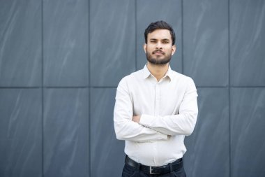Çapraz kollu genç işadamı modern bir ofis binası önünde poz veriyor, şirket ortamında güven, başarı ve liderliği temsil ediyor.
