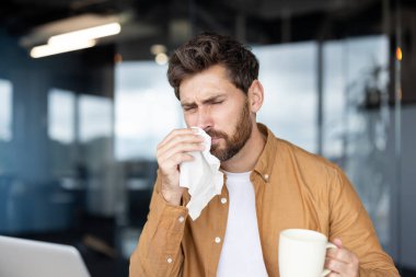 Ofiste dizüstü bilgisayarla çalışırken soğuk algınlığı ya da grip belirtileri gösteren bir işadamı burnuna kağıt mendil sümkürerek diğer elinde bir kupa tutuyor.