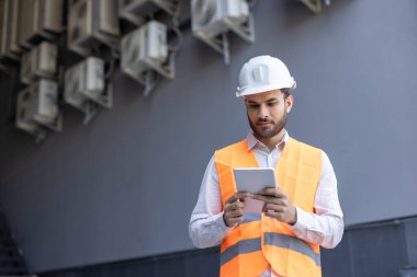 Bina bakım teknisyeni güvenlik kaskı ve yeleği giyiyor, dijital tablet kullanıyor ve dışarıdaki havalandırma ünitelerini denetliyor.