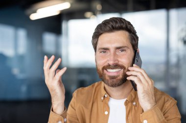 Cep telefonuyla konuşan, modern ofiste olumlu iş konuşmalarının keyfini çıkaran, başarı ve memnuniyeti ifade eden sakallı girişimci.