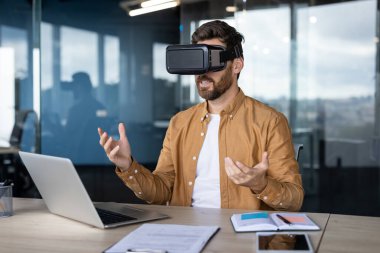 VR kulaklık kullanan ofis çalışanı, modern ofisteki sanal gerçeklikle etkileşime geçiyor. Elleriyle el kol hareketi yaparak, dizüstü bilgisayarla ve belgelerle oturuyor.