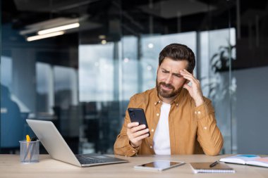 Stresli iş adamı, akıllı telefonundan rahatsız edici haberler okurken alnına dokunuyor, şık, modern ofis ortamında masasında oturuyor, bunalmış ve endişeli hissediyor.