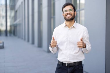 Modern ofis binasının yanında duran mutlu işadamı, iş hayatında olumlu ve başarılı olduğunu belirterek iyi niyet gösteriyor.