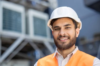 Kask ve güvenlik yeleği giyen Hintli mühendis, bir sanayi ortamında, muhtemelen bir depo veya fabrikada, arka planda HVC birimleriyle güvenle gülümsüyor.
