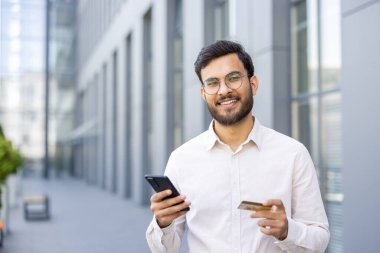 Hintli işadamı elinde akıllı telefon ve kredi kartı tutarken gülümsüyor. Modern bir ofis binasının dışında online işlem yaptığını gösteriyor.