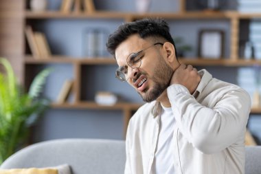 Acı dolu bir ifadeyle boynunu tutan adam, rahatsızlık ve strese işaret ediyor. Oturma odasındaki kanepede oturmak, fiziksel acı ya da yaralanma konseptini aktarmak, muhtemelen duruş ya da stres yüzünden..