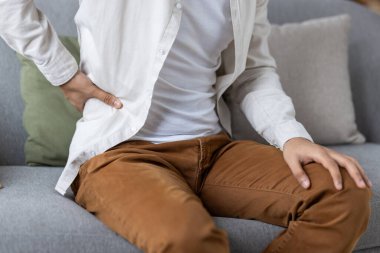 Kanepede oturan genç adam sırt ağrısı çekiyor. Günlük giysiler içinde, sırtı ağrılı bir ifadeyle aşağıda duruyor. Sahne, duruş veya gerginlikle ilgili rahatsızlığı veya potansiyel sağlık sorununu gösteriyor.