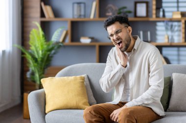 Diş ağrısı çeken genç bir adam çenesini kanepede tutuyor. Ağrıyı, diş sorunlarını ve rahatsızlığı ifade eder. Çağdaş ev kitapları, bitkiler ve yastıklarla iç mekanlar rahat bir atmosfer yaratıyor..