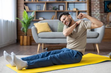 Adam yoga minderinde oturup kolunu esnetiyor. Oturma odasında omzunun ağrıdığını açıkça hissediyor. Kapalı alanda egzersiz sahnesi, fiziksel aktivite sırasında rahatsızlığa ve sakatlığa vurgu yapılır.