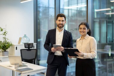 Gülümseyen iki işadamı, elinde dijital bir tablet ve dokümanlarla, geniş pencereli, takım çalışması ve profesyonelliği sergileyen modern bir ofiste duruyorlar.