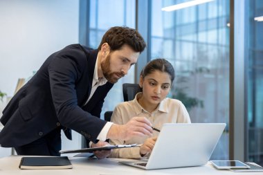 İki iş arkadaşı bir dizüstü bilgisayarda birlikte çalışıyorlar, modern ofis ortamında bir projeyi tartışıyorlar, takım çalışması ve işbirliği gösteriyorlar.