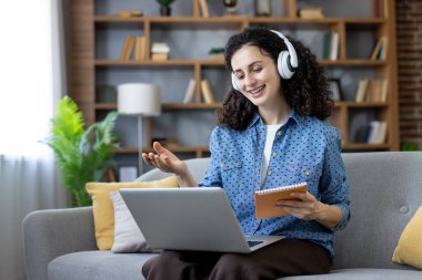 Kulaklık takan ve elinde not defteri tutan mutlu bir kadın, online toplantıda rahat bir oturma odasında kanepede otururken dizüstü bilgisayar kullanarak konuşuyor..