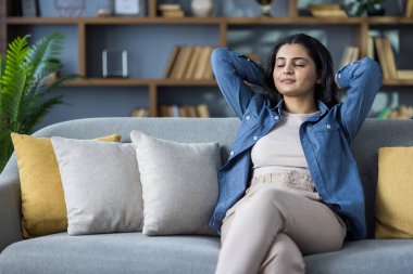 Relaxed young Indian woman resting on the couch at home, sitting with her eyes closed and her hands behind her head.