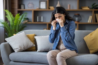 Sick young Indian woman sits on the couch at home and wipes her nose with a tissue from a runny nose.