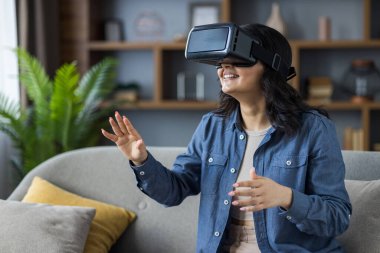 Indian young smiling girl sitting on sofa at home wearing virtual reality glasses and gesturing with hands, having fun online.