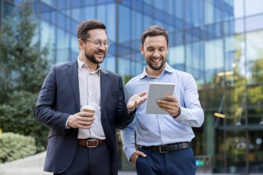 Two cheerful male colleagues brainstorming and collaborating on a new business project using a digital tablet while standing outdoors in front of a modern office building
