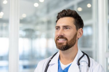 Smiling male doctor in white lab coat with stethoscope, confident and approachable, working in a bright modern clinic setting professional, caring, and trustworthy clinician