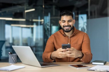 Young indian businessman sitting at his desk in a contemporary office, happily navigating on his smartphone, representing concepts like mobile communication, business technology, and connectivity