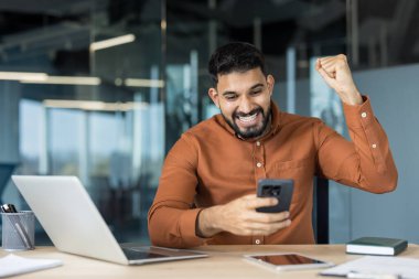 Happy bearded man sitting at a desk in a modern office, raising his fist in celebration while reading exciting news on his mobile phone, experiencing success and achievement