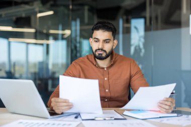 Young man analyzing financial reports and paperwork, using a laptop, and working diligently in a modern office environment, focusing on professional tasks