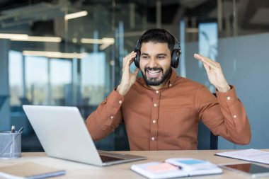 Happy young man wearing headphones, talking and gesturing during an online video conference or webinar on a laptop in a modern office, enjoying remote communication