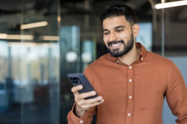Young indian businessman smiling broadly while holding and looking at a smartphone, engaged in digital communication, online work, or social media in a modern office environment