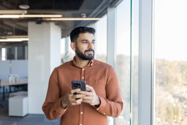Young professional man standing in a modern office, holding his smartphone and thoughtfully looking out through a large window, contemplating new business opportunities and future plans