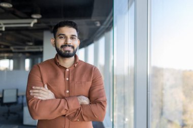 Young indian businessman with a beard and a warm smile, wearing a brown shirt, standing confidently with arms crossed in a contemporary office space next to a large window