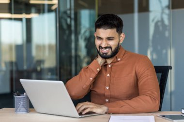 Young businessman experiencing severe neck pain and discomfort while working long hours on his laptop in a modern office, highlighting work-related stress and poor ergonomics
