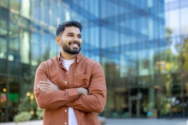 Young indian businessman smiling confidently with arms crossed, standing outdoors in front of a modern glass office building, representing corporate success and professional urban lifestyle