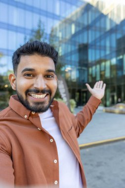 Young man smiling at camera, making a selfie video for social media and pointing to a contemporary glass office building in an urban business district