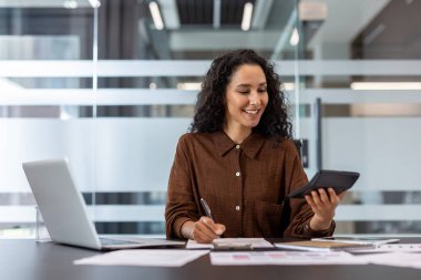 Young businesswoman smiling, calculating financial data with a calculator, writing notes, and working at her desk in a bright, modern office environment