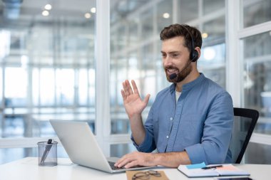 Man wearing an ear-hook headset while smiling and waving during a video call on his laptop in a contemporary bright office, representing remote work and communication