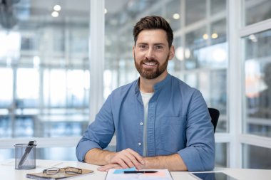 Smiling bearded man sitting at a desk in a bright, modern office, looking confident and experienced while working in a professional, comfortable setting