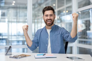 Happy businessman cheering with raised fists, celebrating achievement and triumph while working at a modern office desk, embodying success, motivation, and positive career growth